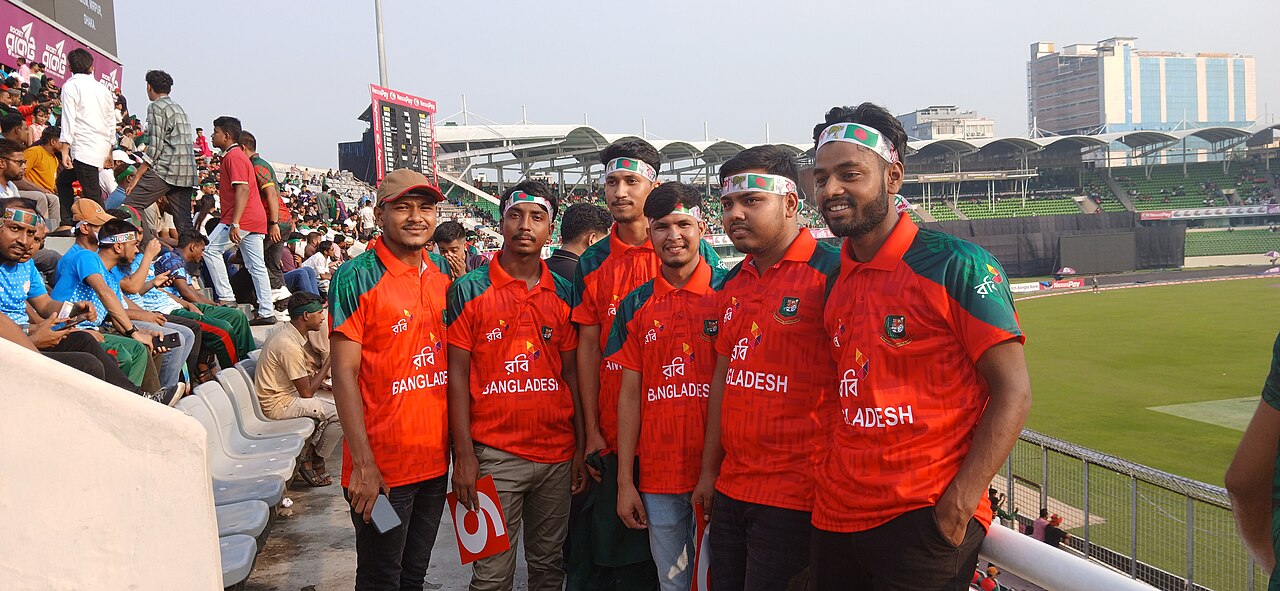 Spectators in the stands of a cricket stadium wearing green Pakistan shirts, with green flags and a Bangladesh flag visible in the seating area