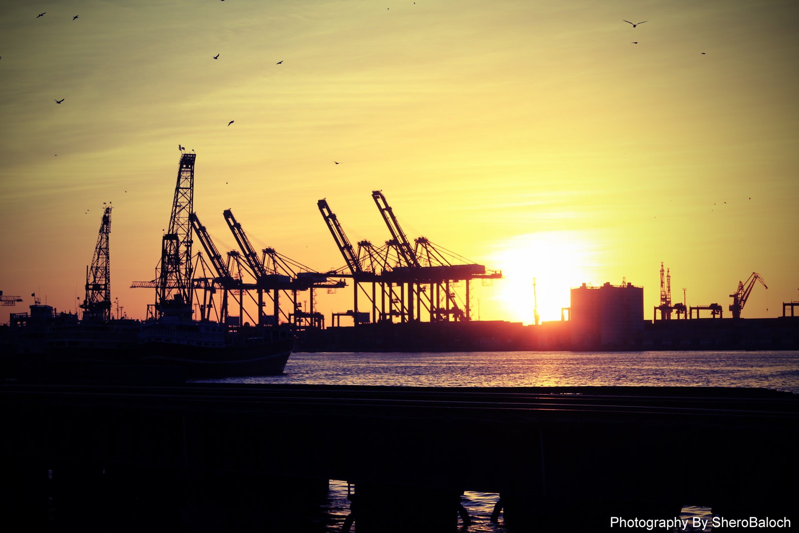 Silhouetted gantry cranes and a berthed cargo ship at Karachi Port against a golden sunset, with a warehouse and harbour buildings along the waterfront.