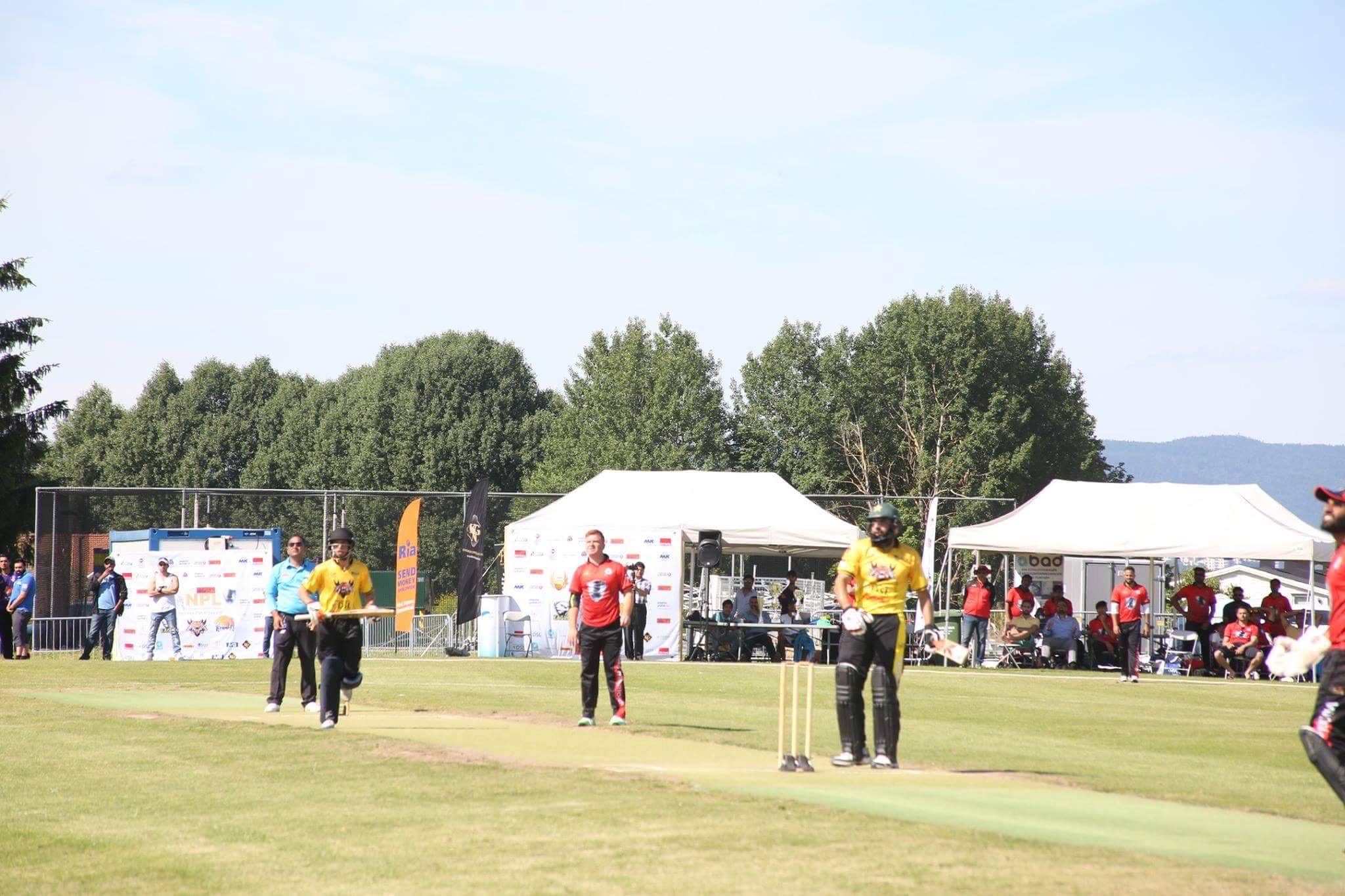 A right-handed batter in dark cricket gear plays a shot in the middle of a green outfield while the wicketkeeper and slip fielders watch the ball, with a bank of spectators visible behind a boundary rope