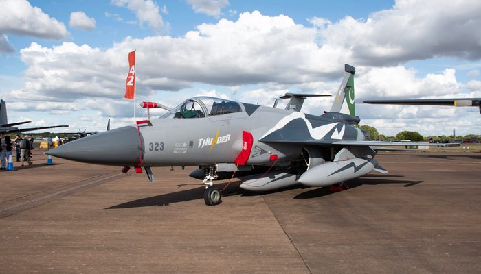 A grey Pakistan Air Force JF-17 Thunder fighter jet marked 'THUNDER' and numbered 323, parked on a tarmac under a partly cloudy sky with an orange flag on a pole beside it.