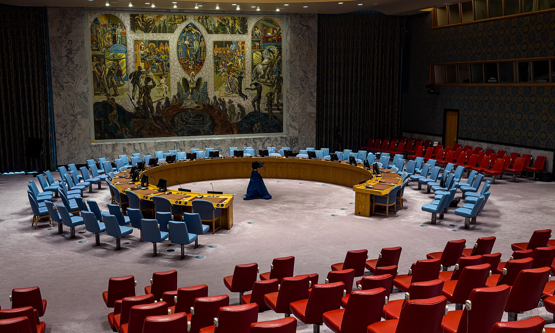 The empty United Nations Security Council chamber in New York, with blue delegate chairs around a curved wooden table, red gallery seats in the foreground and a large mural on the far wall.