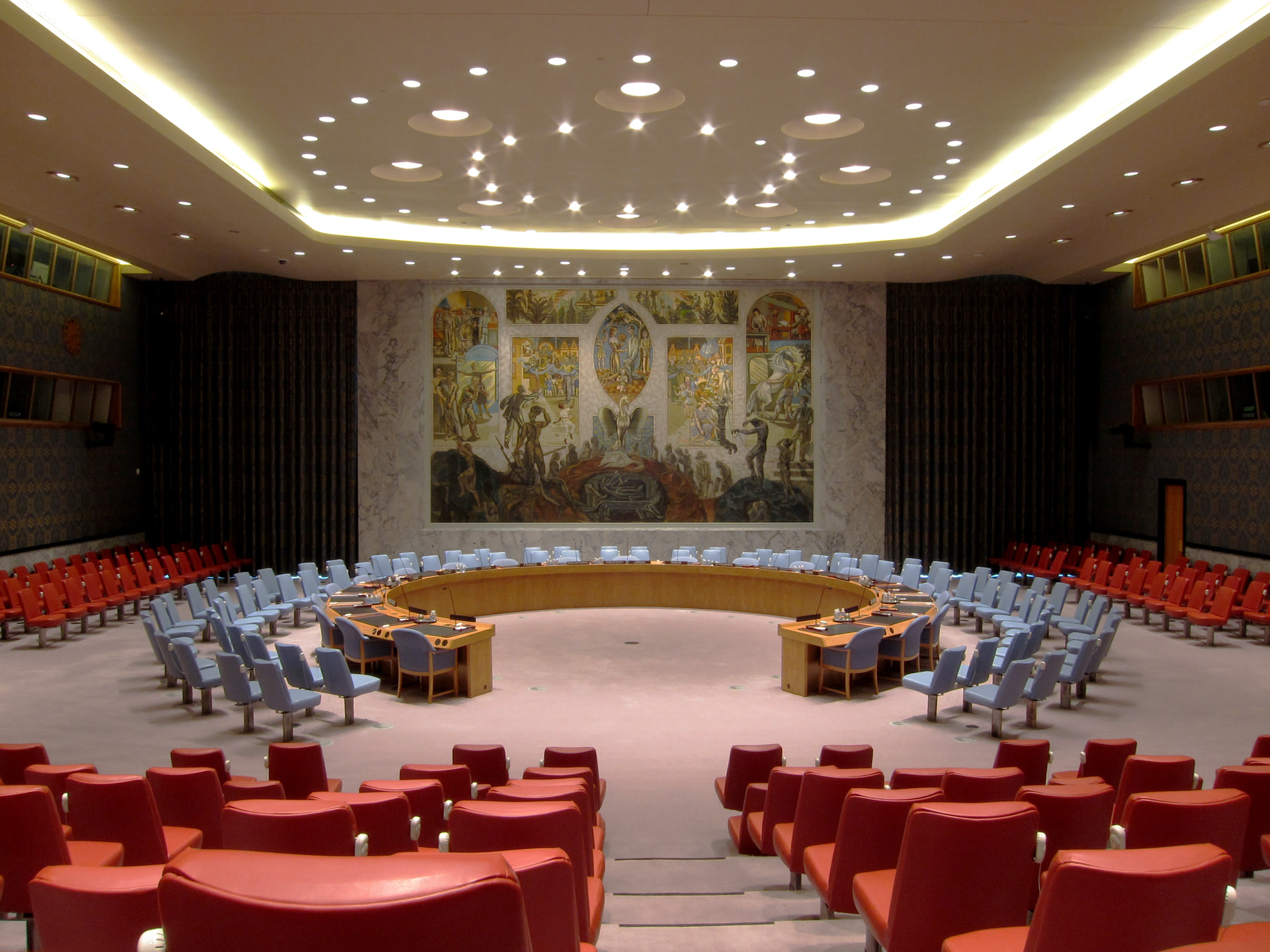 Wide view of the United Nations Security Council chamber in New York, showing the horseshoe-shaped meeting table and wall mural