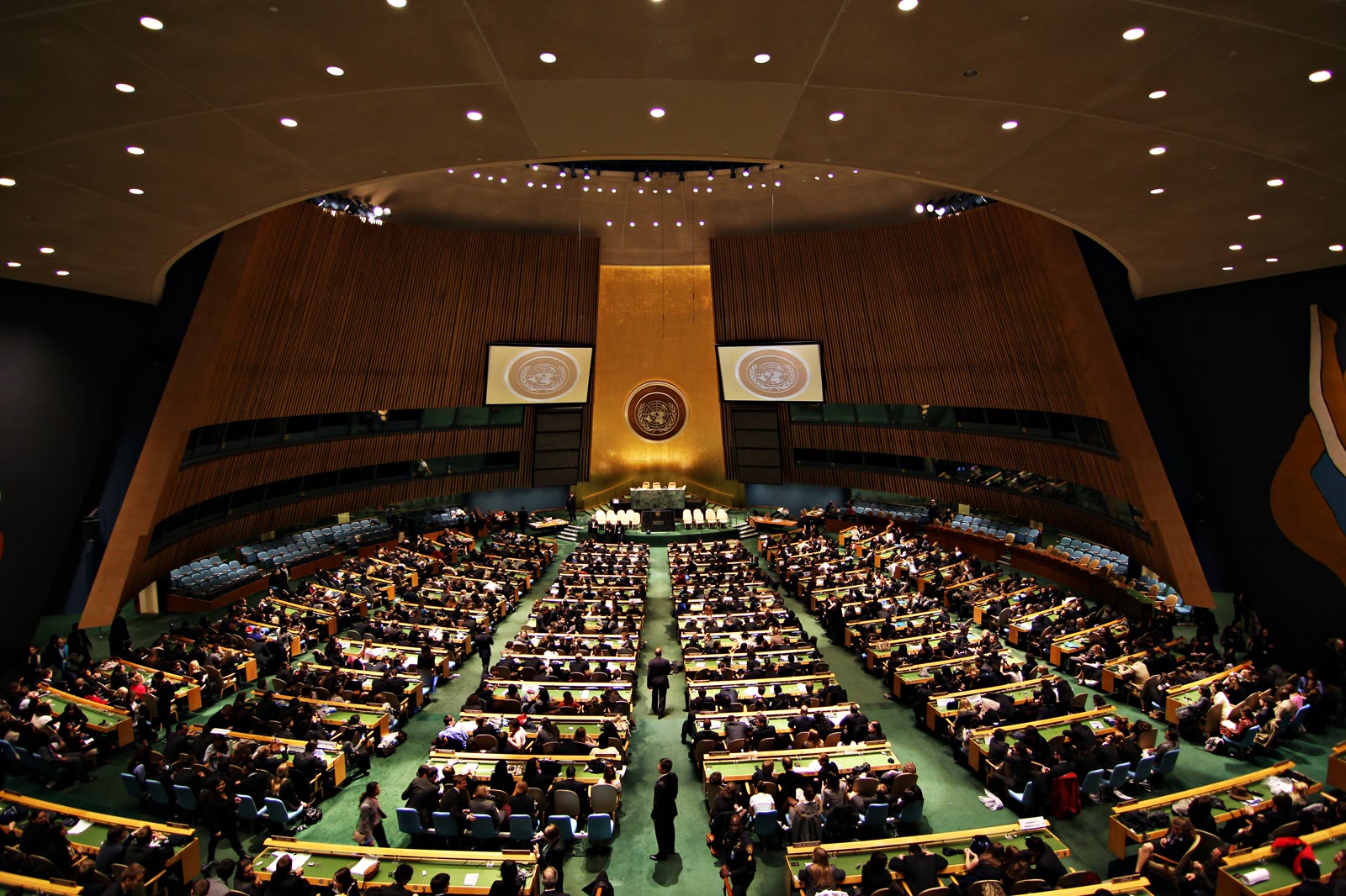 The General Assembly hall at UN Headquarters in New York, seen from the upper gallery, with the gold United Nations emblem above the podium, two screens either side of it and rows of green desks filled with delegates.