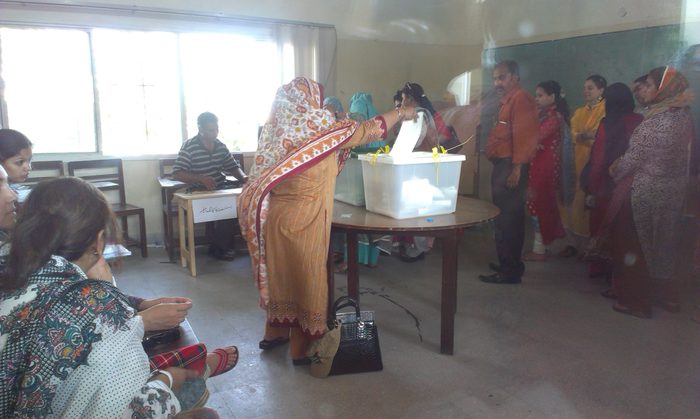 Women voters at a polling station, one in a patterned shawl placing a paper ballot into a white box on a table while others wait their turn.