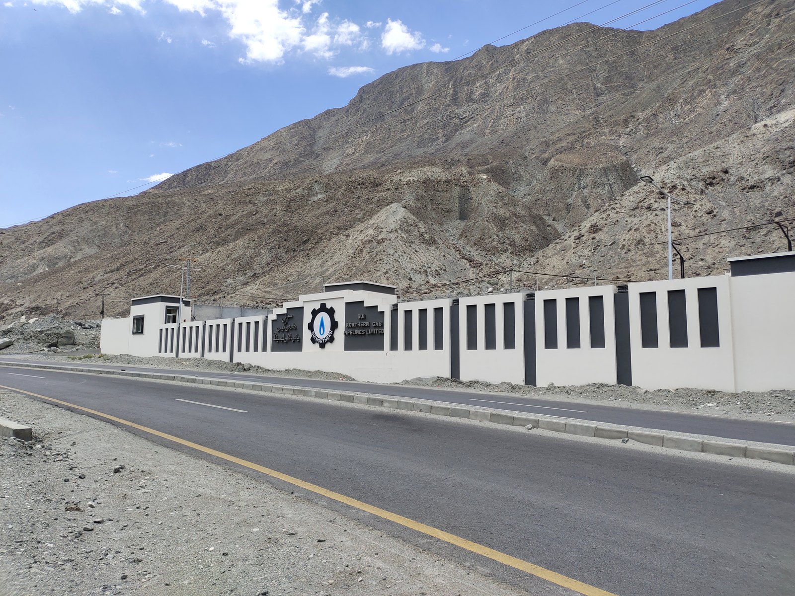 The entrance wall and gate of the Sui Northern Gas Pipelines plant in Gilgit, with the company name in English and Urdu on black panels, street lights and overhead lines along an empty road, and a bare brown mountain slope rising behind.