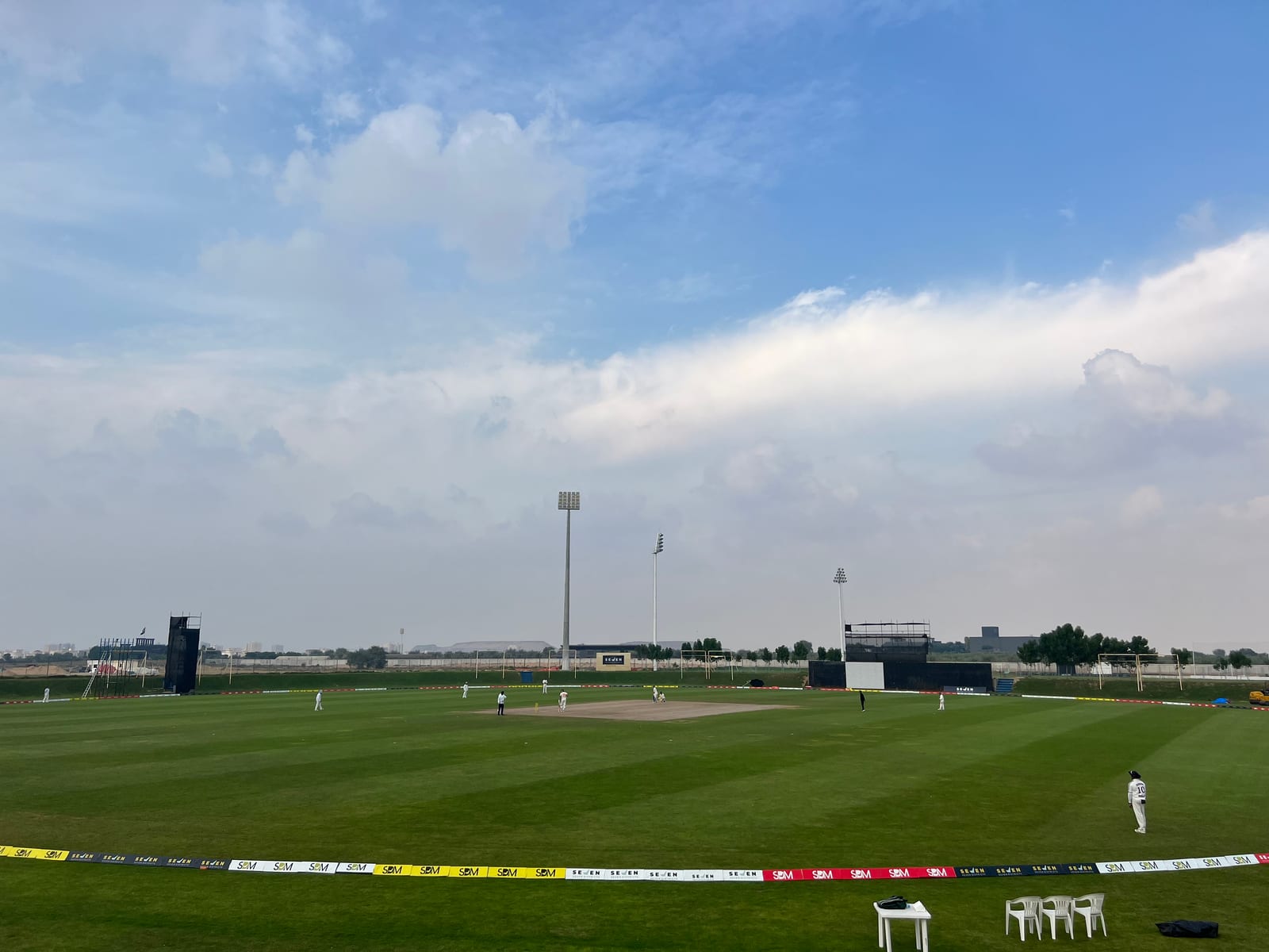 A cricket field in Ajman with floodlights, advertising boards along the boundary and fielders spread across the outfield under a partly cloudy sky