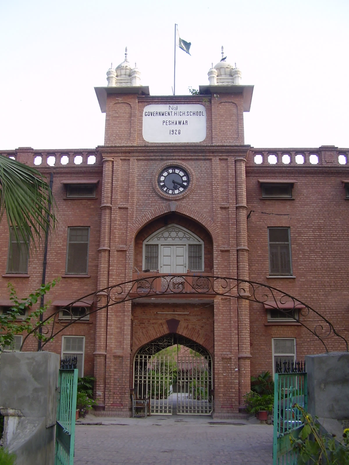 The façade and gated entrance of a Peshawar government school building, with a tall flagpole, three stories of classrooms, and mature trees shading the courtyard.
