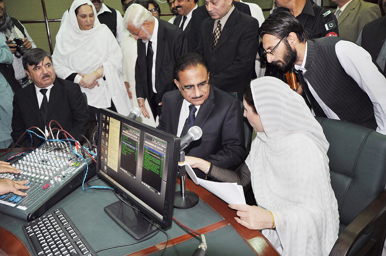 The Chief Justice of the Peshawar High Court inaugurating the Khyber Pakhtunkhwa Judicial Academy's radio station, with officials seated behind a microphone.