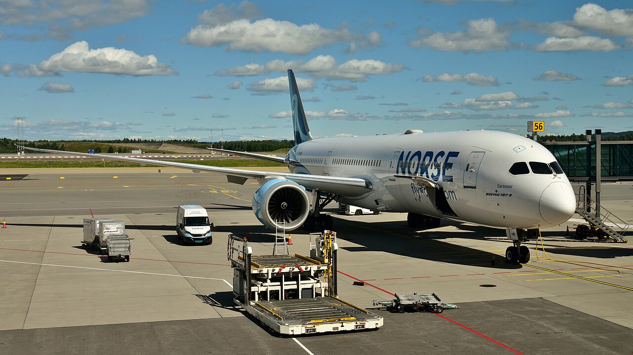 A Norse Atlantic Airways Boeing 787-9 in white and blue livery parked at an airport gate with a jet bridge attached, support vehicles on the apron, blue sky and scattered clouds in the background.