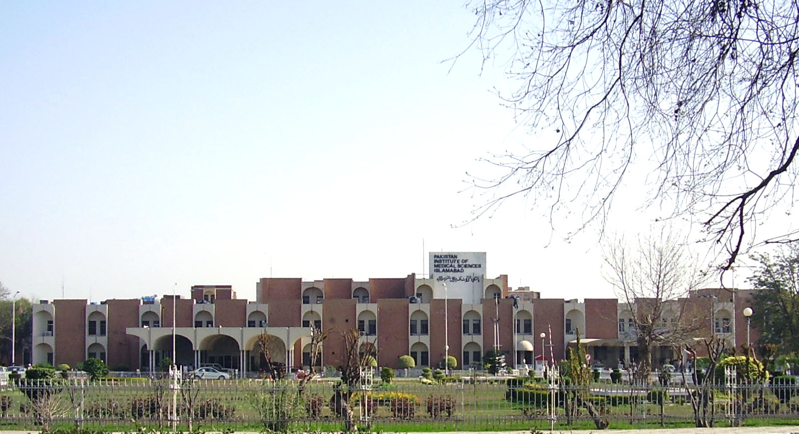 A multi-storey hospital building with a pale facade and rows of windows seen across an open paved area under a clear sky.