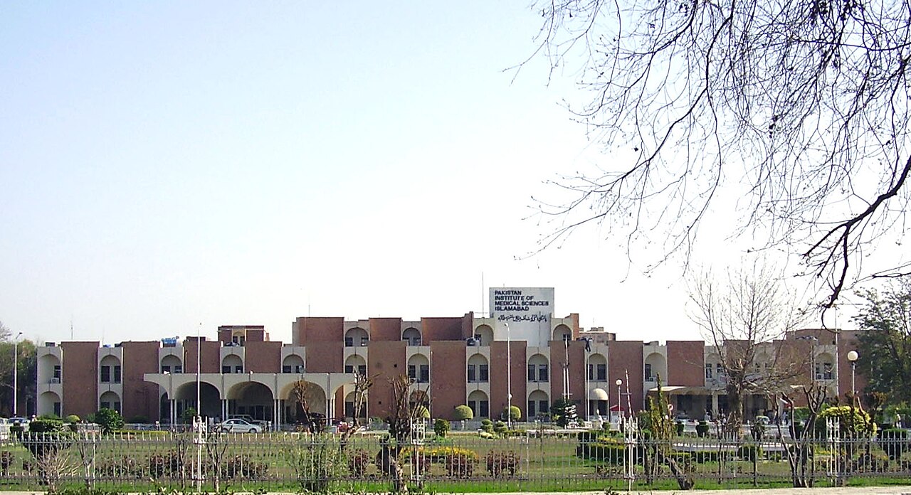 The red-brick main building of the Pakistan Institute of Medical Sciences seen across a lawn, with its name on a rooftop sign.