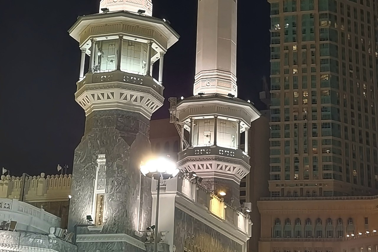 The exterior of the Grand Mosque of Makkah in Saudi Arabia seen at night, with the clock tower and surrounding buildings lit up.