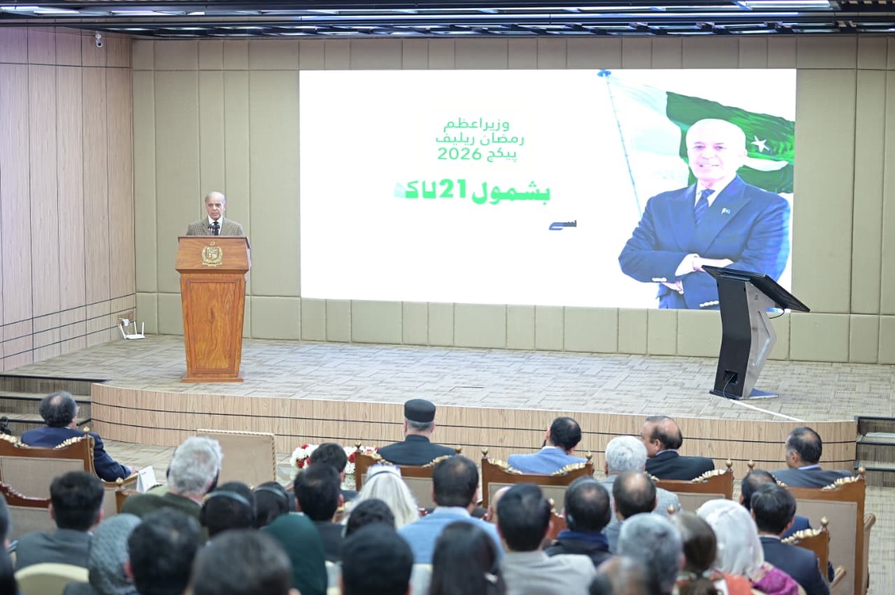 Prime Minister Shehbaz Sharif stands at a wooden podium during the launch ceremony of the Ramazan relief package as a large screen displays the programme's title and his portrait.