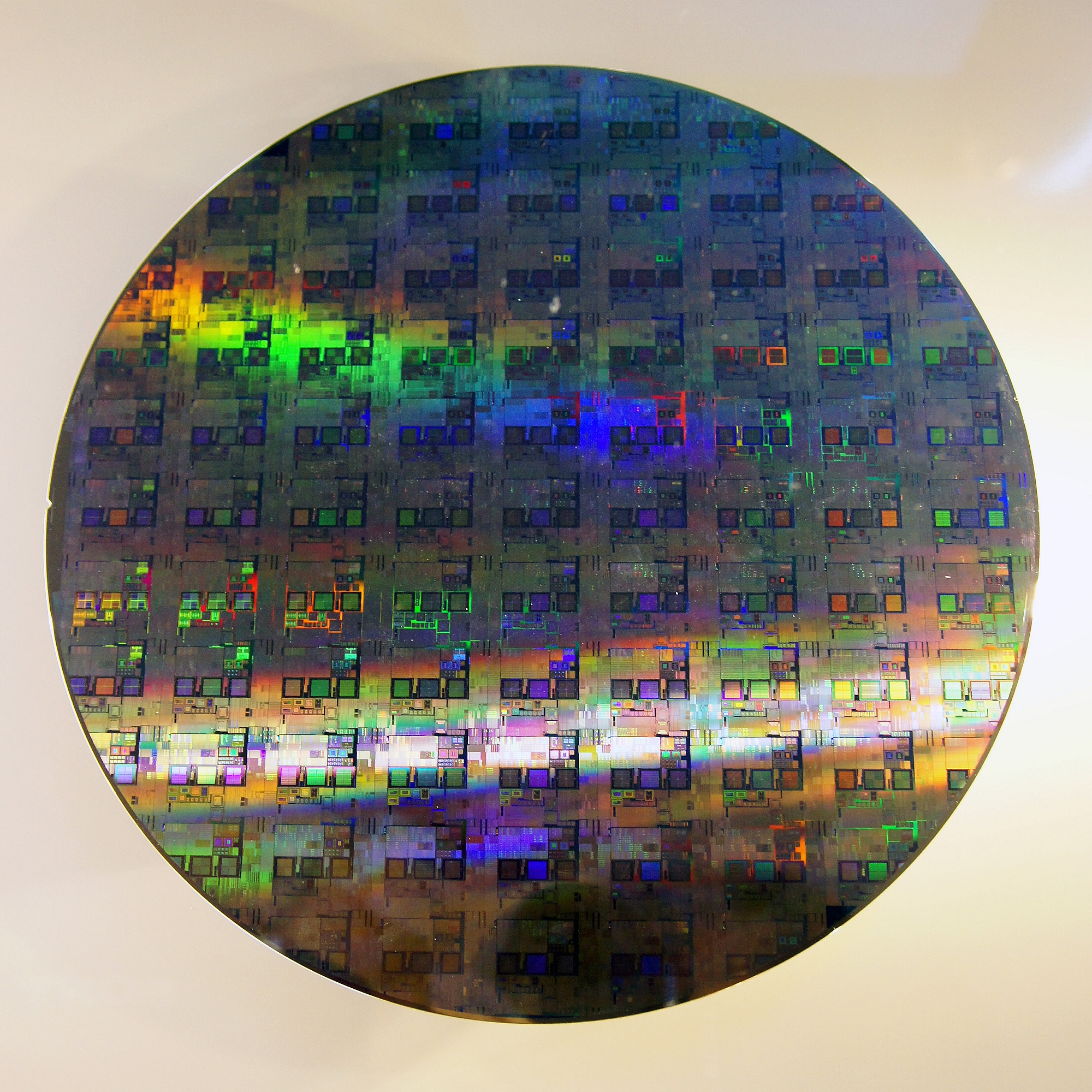 A circular 12-inch silicon wafer reflecting iridescent rainbow colours, the substrate on which semiconductor chips are fabricated.
