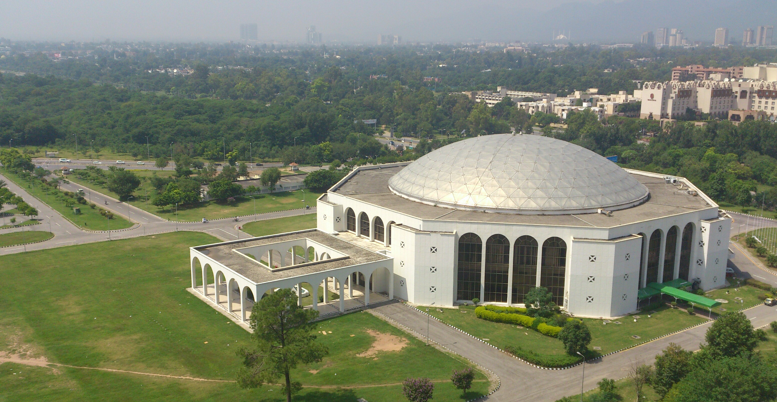 The Jinnah Convention Centre in Islamabad, a long white modernist building behind a flagged entrance plaza, photographed in daylight