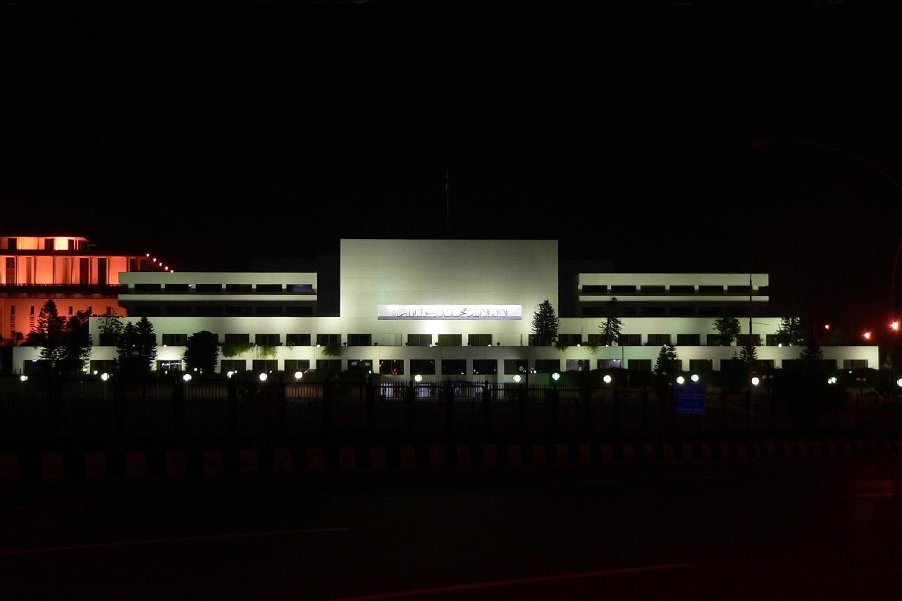 A wide night-time view of the illuminated Pakistan Parliament House in Islamabad, with its central dome flanked by lower wings and reflecting pools in the foreground.