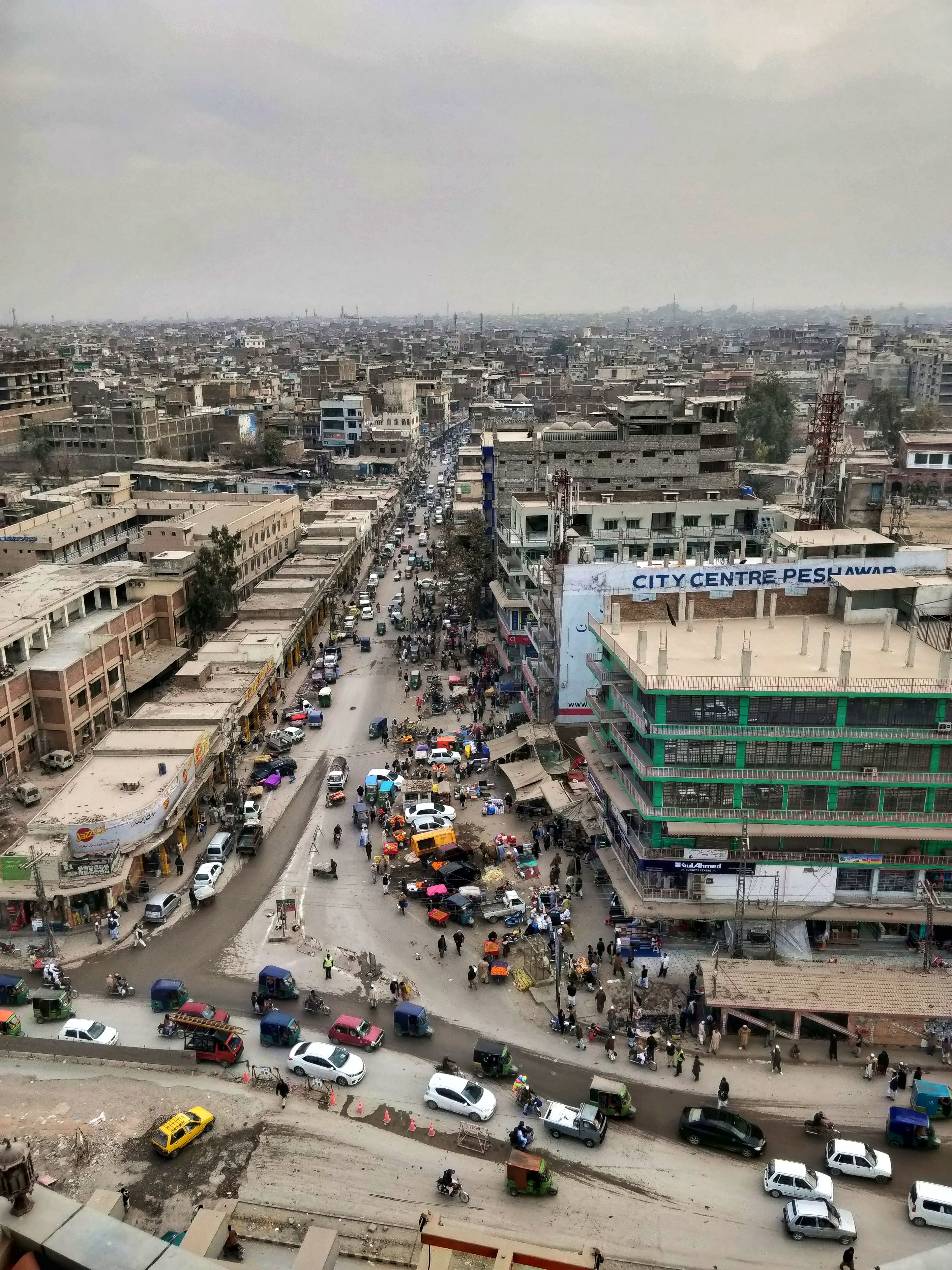 An elevated view over a busy commercial street in central Peshawar, with low-rise shopfronts, parked cars, rickshaws and pedestrians crowding the roadway.