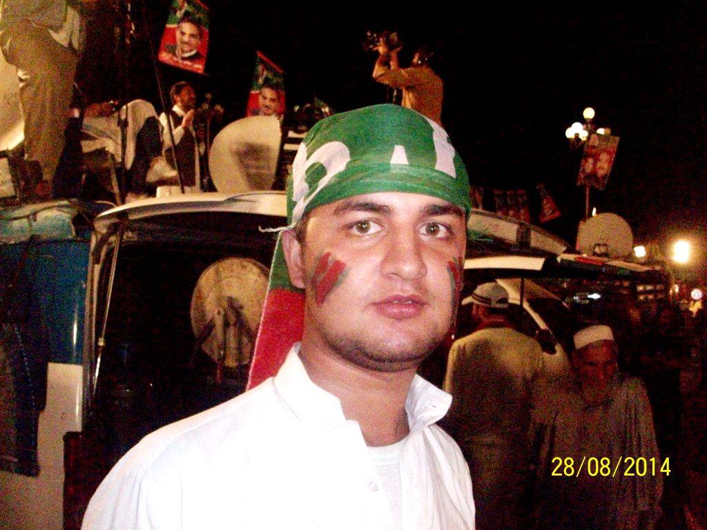 A man with red-and-green face paint and a green PTI flag wrapped around his head stands in a night-time rally crowd, with flags and framed portraits behind him.