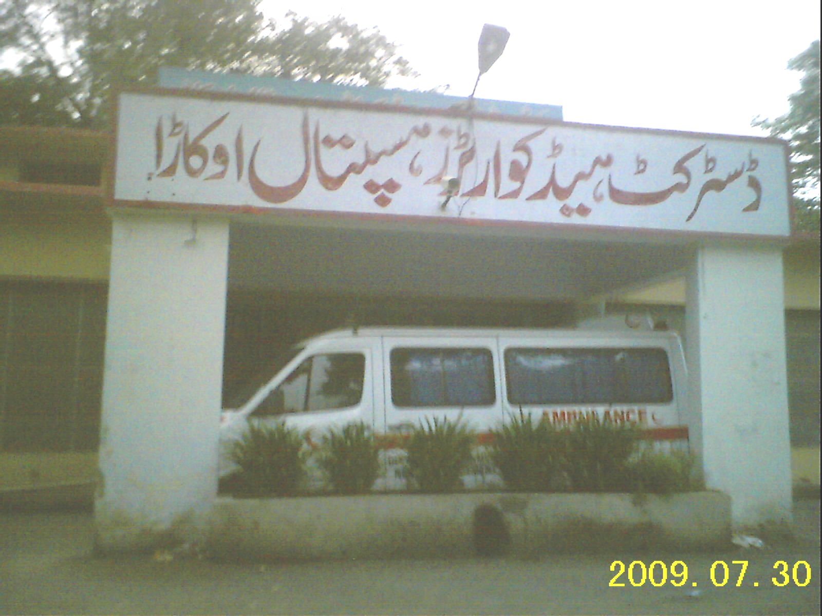 The exterior of a district headquarter hospital building in Pakistan, showing an ambulance parked in front of the old, single-storey medical facility