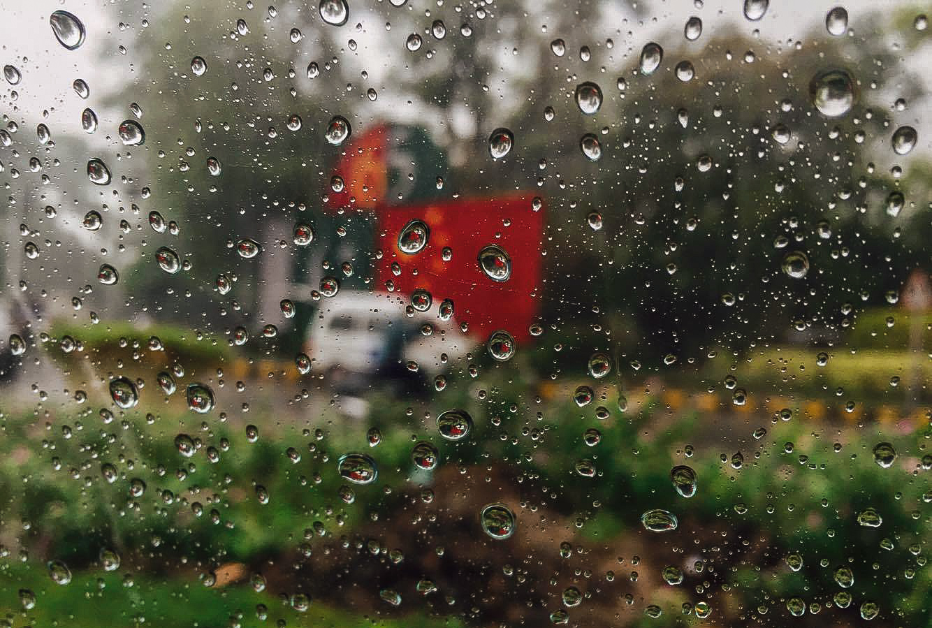 A rain-soaked street in Lahore under an overcast sky, with water on the ground and low buildings along the roadside.