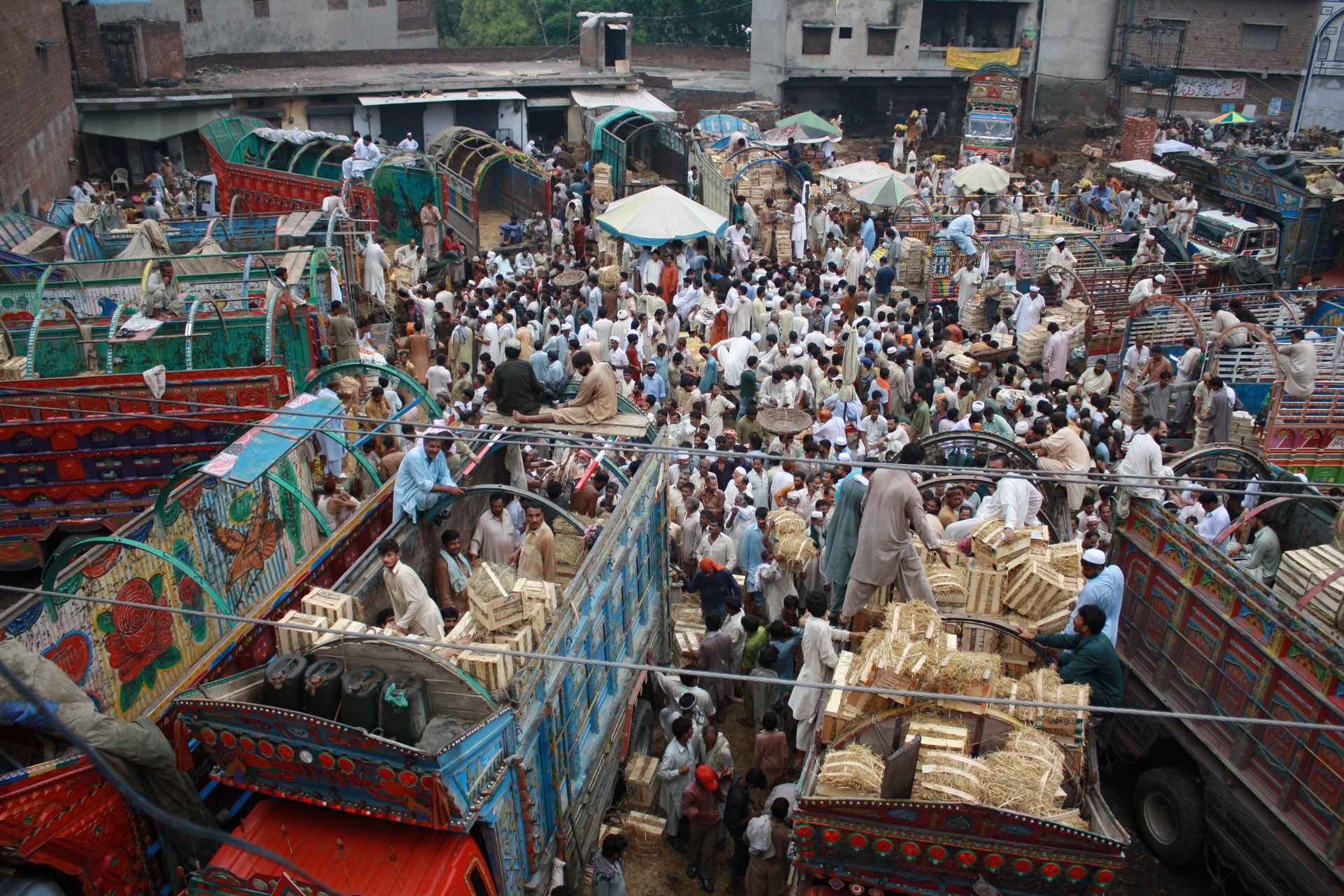 Overhead view of a crowded wholesale fruit market, with traders and buyers standing among brightly painted delivery trucks loaded with stacked crates of produce and bundles of straw.