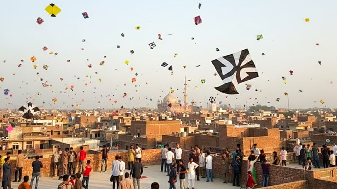 Crowds stand on rooftops in Lahore as dozens of kites fly over the city skyline, with a domed mosque and minarets visible in the distance.