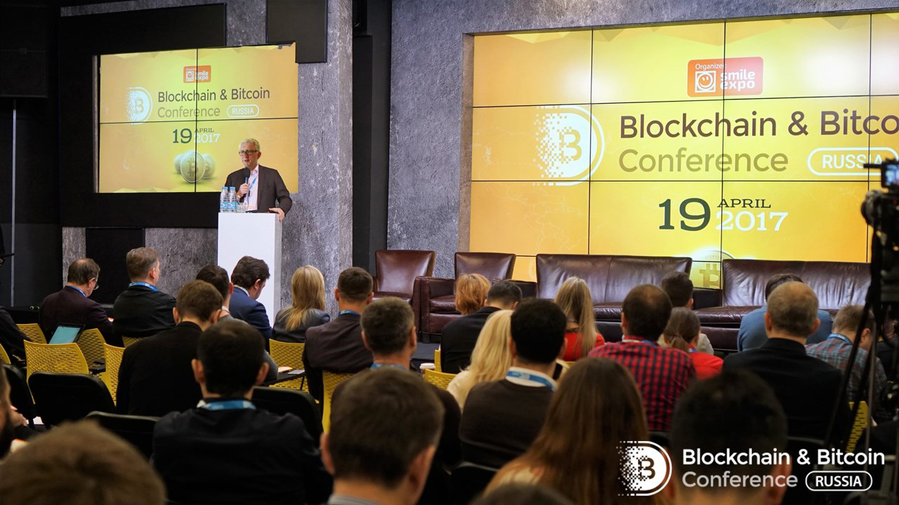 An audience seated in a darkened conference hall looking at a brightly lit stage during a cryptocurrency and blockchain conference, with purple and blue stage lighting and a large presentation screen at the back.