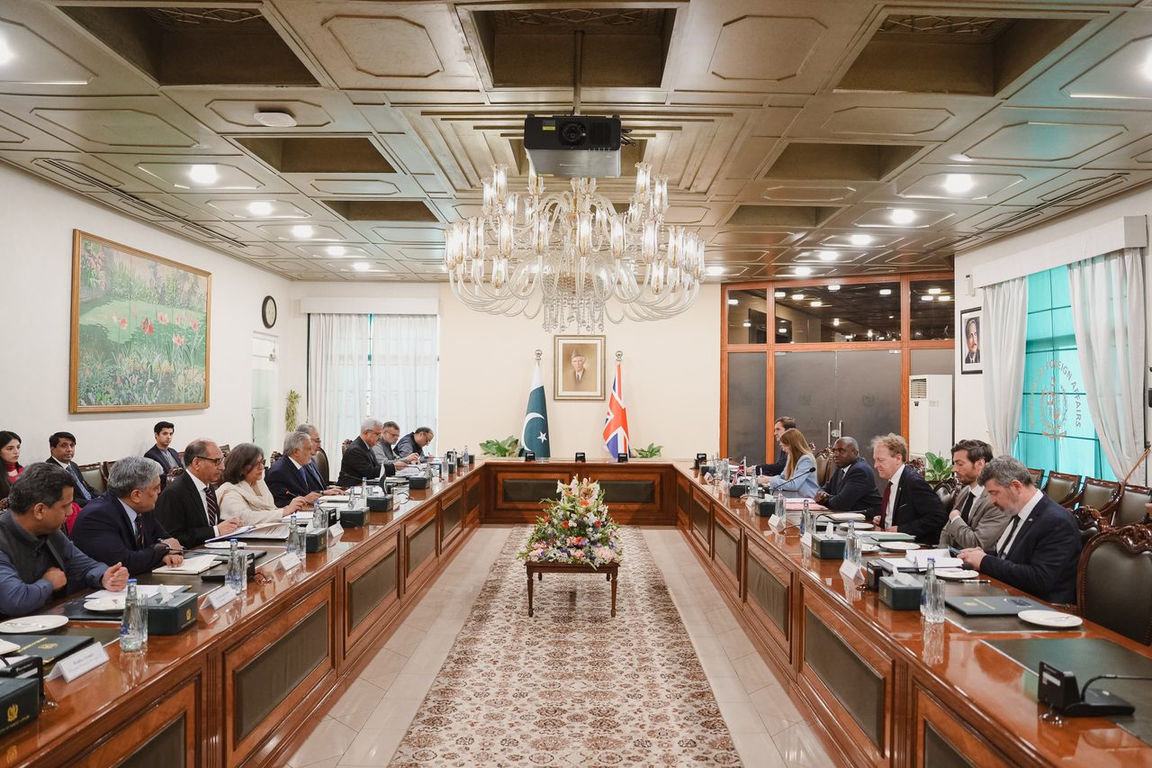 Officials seated along both sides of a long polished conference table beneath a large glass chandelier, with Pakistani and British flags at the far end.