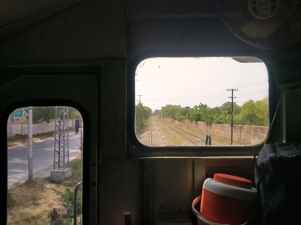 The view from inside a Pakistan Railways locomotive cab, looking along double railway tracks past a roadside wall, trees and a station sign