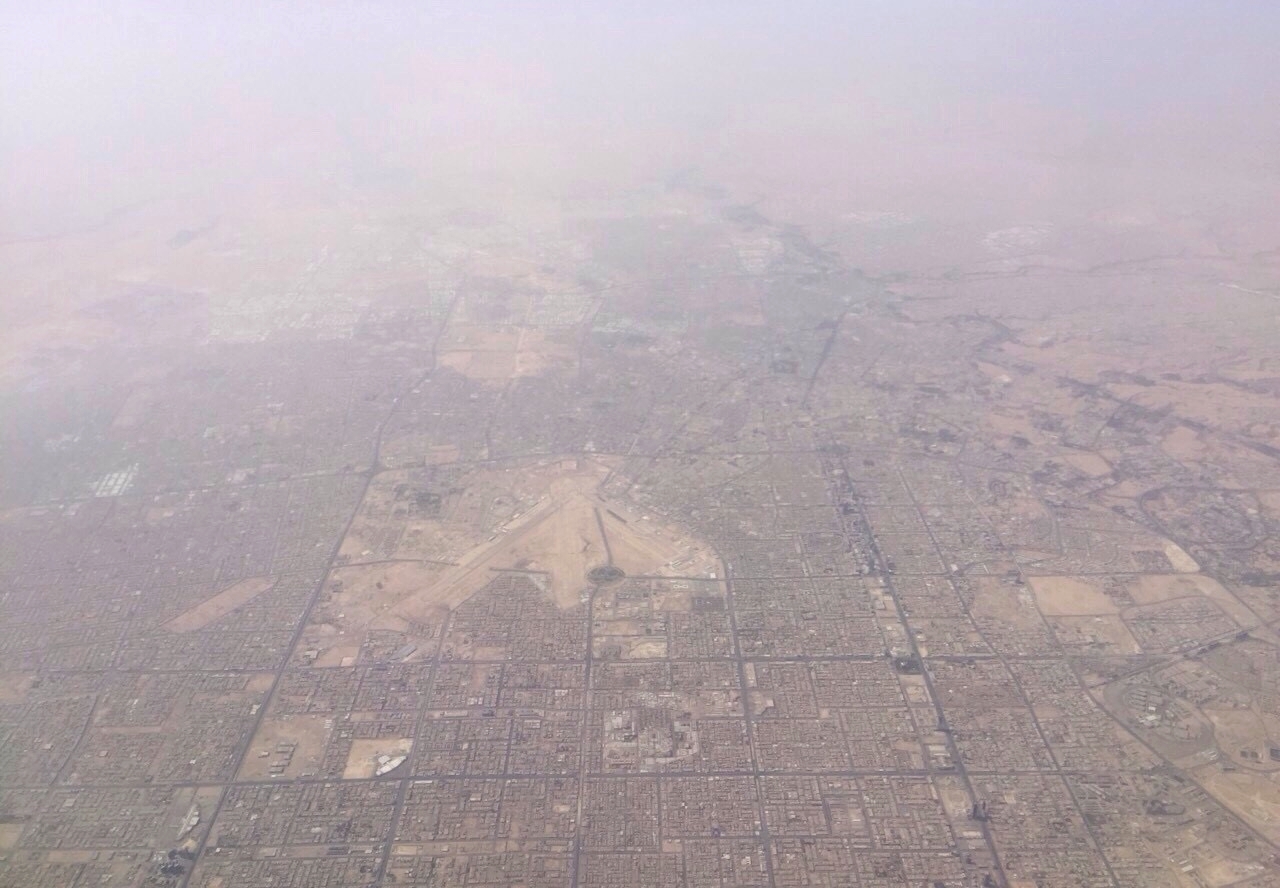 An aerial view over Riyadh showing the terminal buildings and runways of King Khalid International Airport, surrounded by desert terrain.