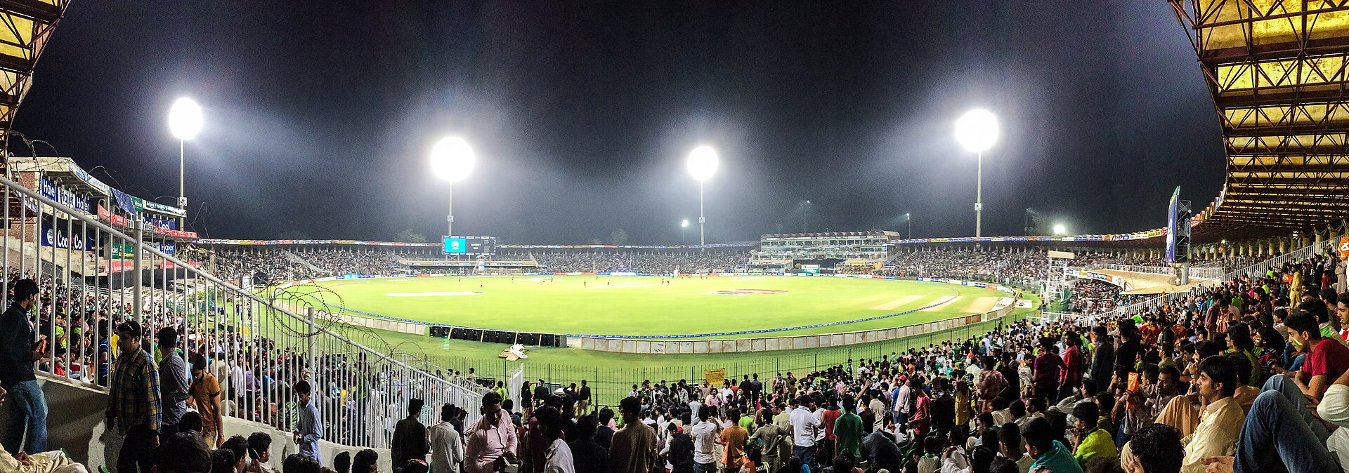 Floodlights over a packed cricket stadium at night, with the green outfield and the square's boundary rope in front of the stands