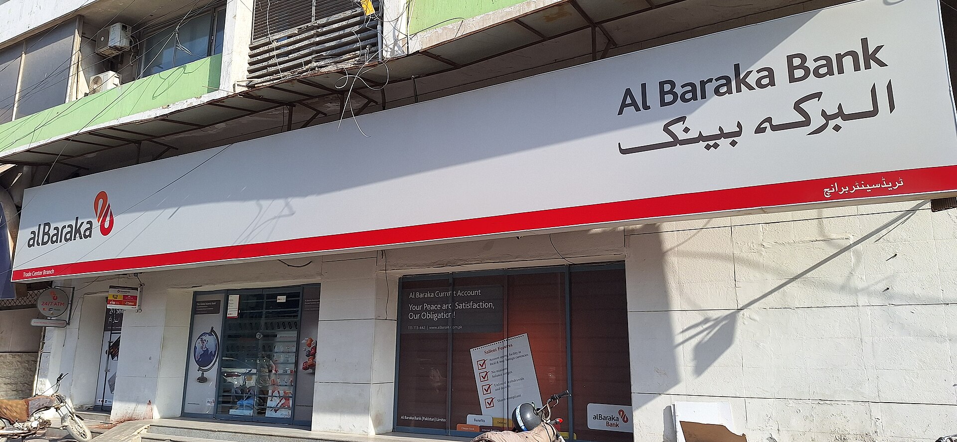 A bank branch frontage on I.I. Chundrigar Road in Karachi, with the bank's name in English and Urdu across a white and red fascia, a 24-hour ATM sign beside the door and a motorbike parked at the kerb.