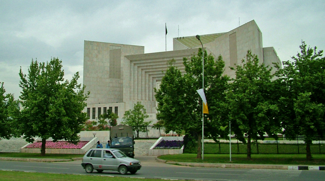 The Supreme Court of Pakistan building in Islamabad, a pale-stone structure with a stepped geometric facade and a flag on the roof, seen across a road with green trees and flower beds in the foreground.