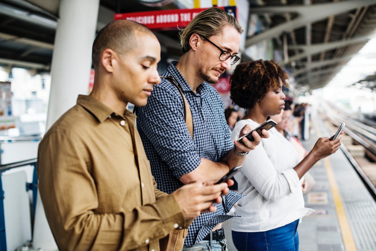 Three people at a railway station, each looking down at a smartphone held in one hand
