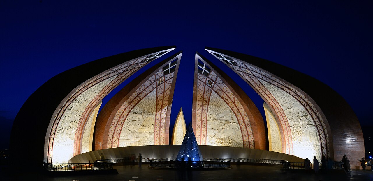 The Pakistan Monument in Islamabad, a large petal-shaped concrete structure at the foot of the Margalla Hills, set against a clear blue sky.