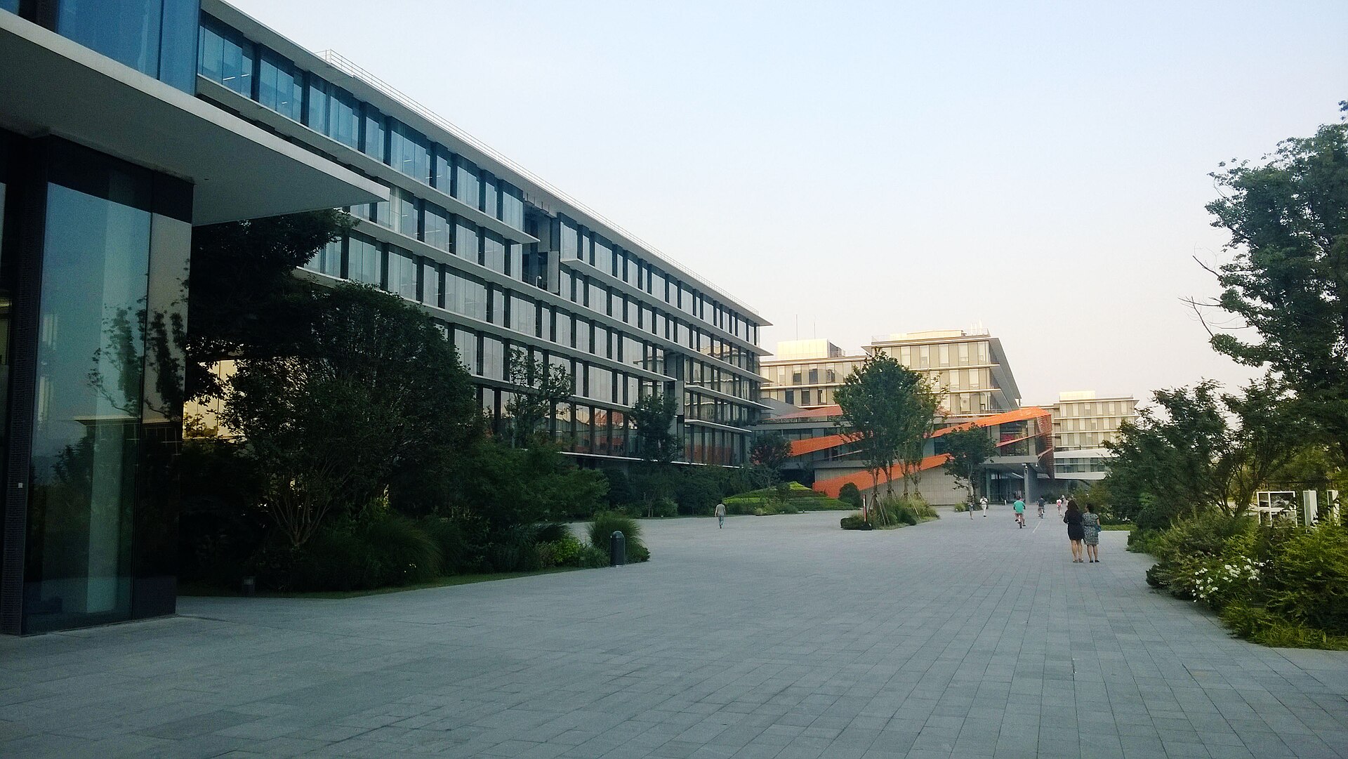 A multi-storey glass office block lining a wide paved plaza at Alibaba Group's headquarters campus in Hangzhou, with mature trees, planted beds and a few people walking across the square.