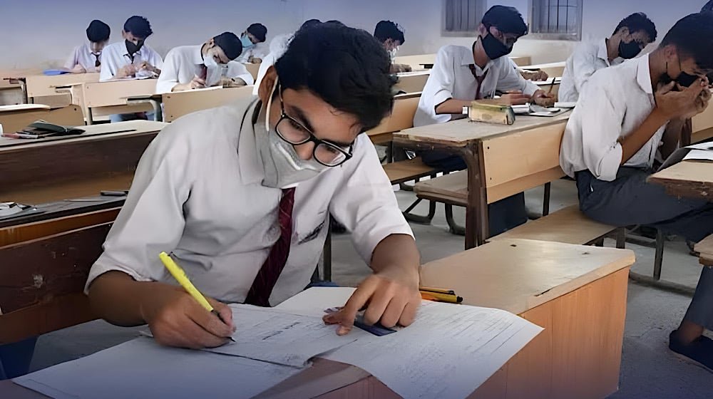 Students in white uniforms and face masks writing an examination in a classroom