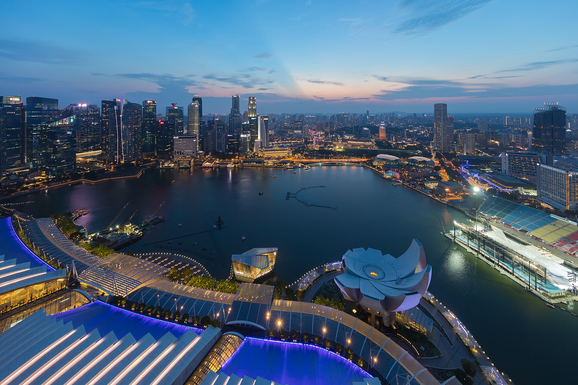 Dusk view across Marina Bay in Singapore from the Marina Bay Sands observation deck, with the lit towers of the central business district across the water, the lotus-shaped ArtScience Museum on the right and the illuminated Supertree Grove on the left.