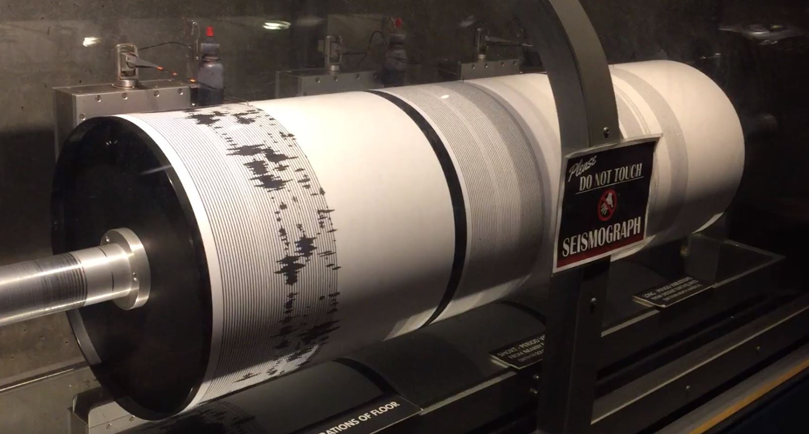 Pen-style seismograph needle tracing dark wavy lines on a rotating paper drum inside a beige instrument cabinet
