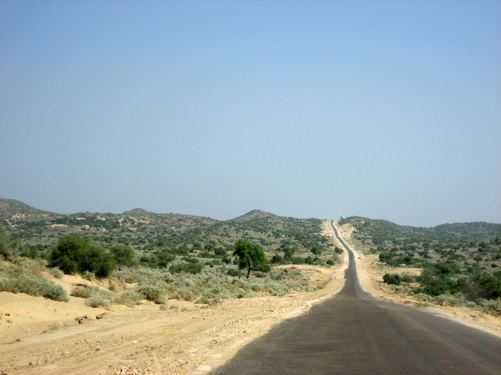 A paved road crossing the arid, scrub-covered landscape of Tharparkar district in Sindh, Pakistan.