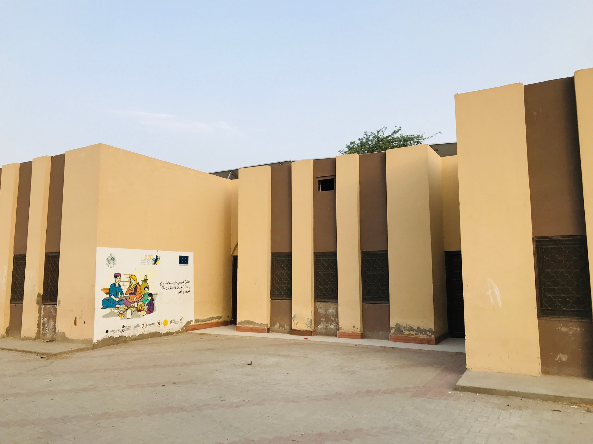 A cream-painted single-storey health centre with tall pilasters, dark metal window grilles and a covered porch, standing behind a wide paved forecourt, with a painted wall mural of a family at a health counter beside Sindh government and aid agency logos.