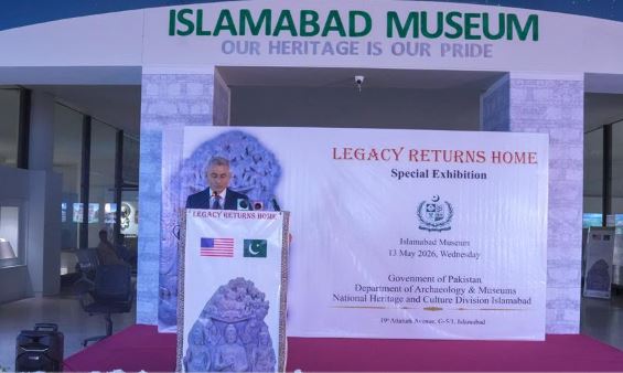 A US official speaks at a podium bearing a 'Legacy Returns Home' exhibition banner, with US and Pakistani flags and the Islamabad Museum sign above.