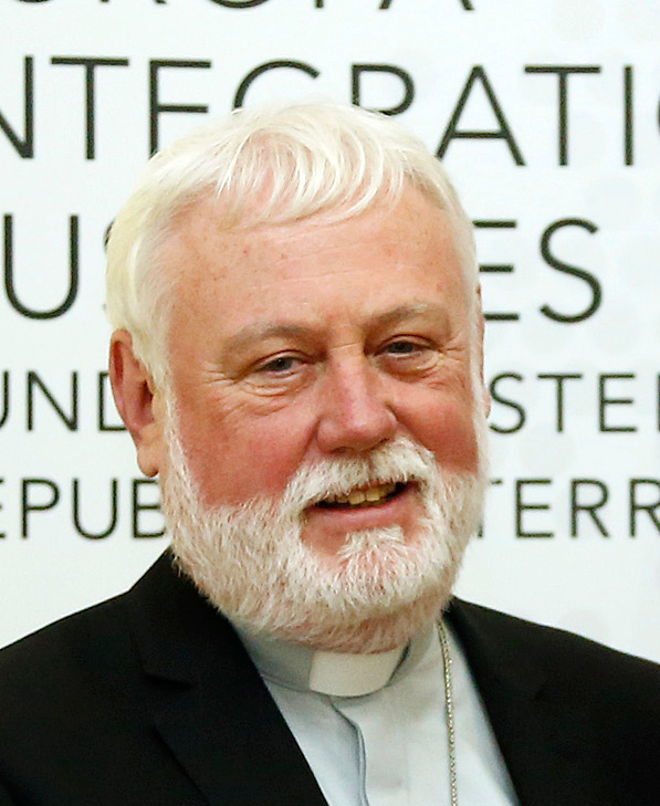 Close-up portrait of Archbishop Paul Richard Gallagher, a white-haired, white-bearded man wearing a dark suit and a clerical collar, photographed indoors in front of a sign.