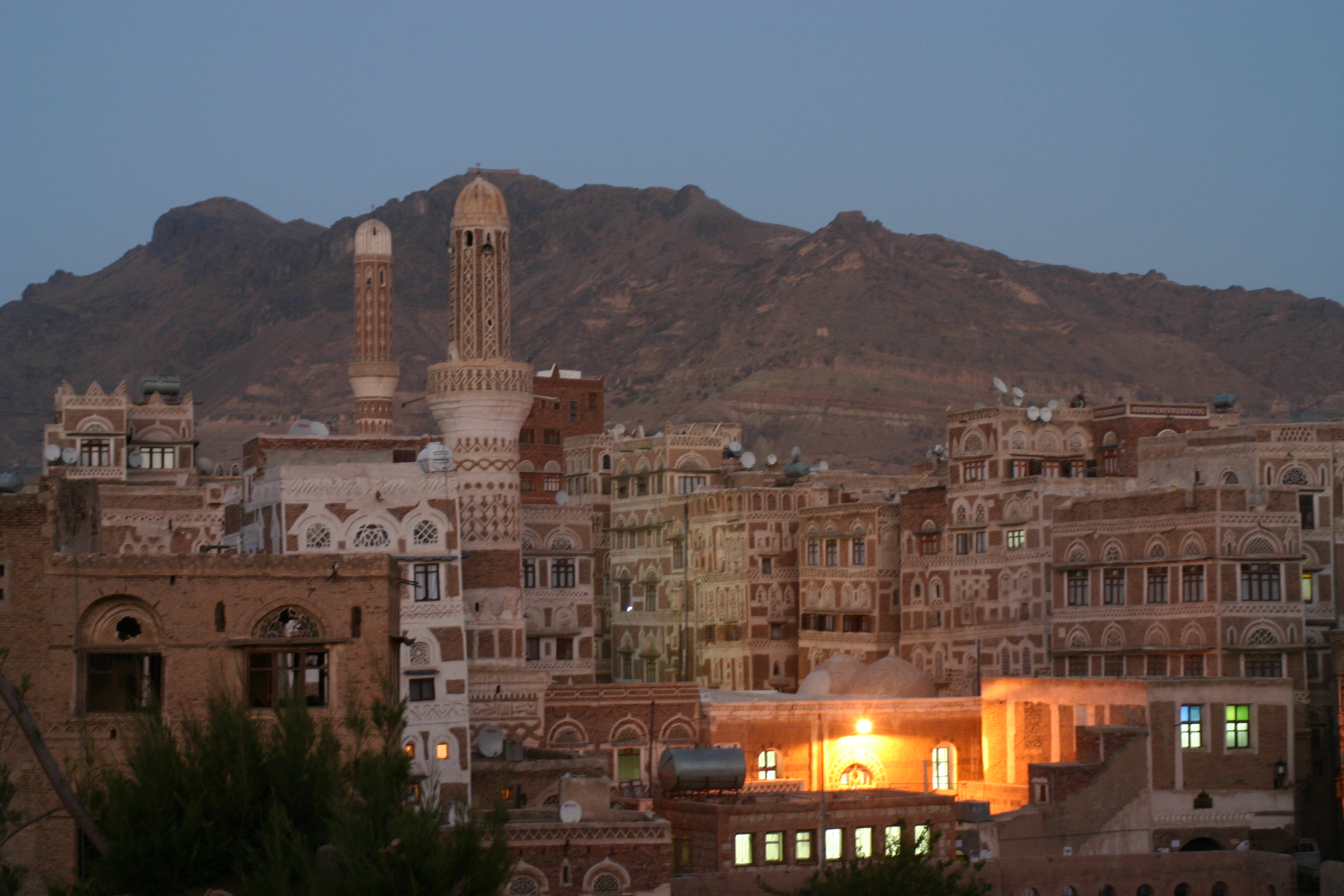 The distinctive mud-brick tower houses of Sanaa's old city skyline at dusk, viewed from an elevated position across the rooftops.